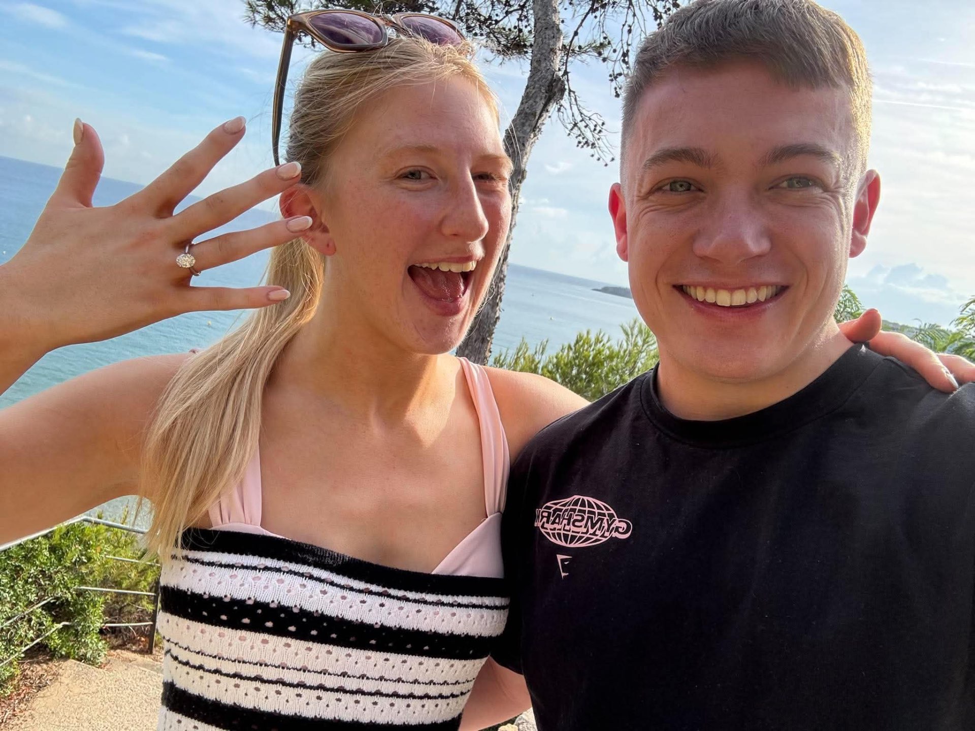 Mollie and Luke smiling together on a coastal path, with the sea behind them